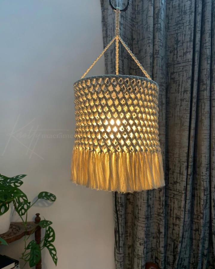 Golden cotton macrame pendant from @knottymacrame_. Diamond knots and long fringe glow warmly beside a leafy plant.