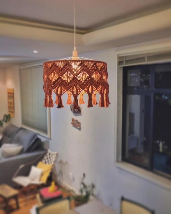 Burnt-orange macrame chandelier from @fuxicandoarteuai. Diamond knots and tassels surround a glowing bulb.