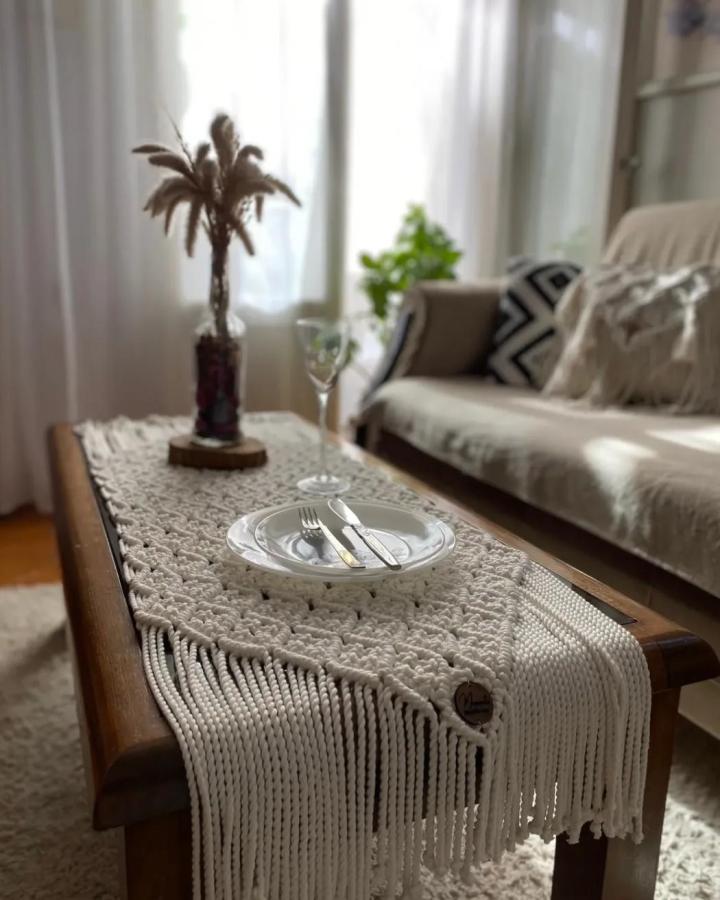 Creamy macrame table runner from @memento.decorandmore. Textured fringe decorates a wooden coffee table in a cozy living room.