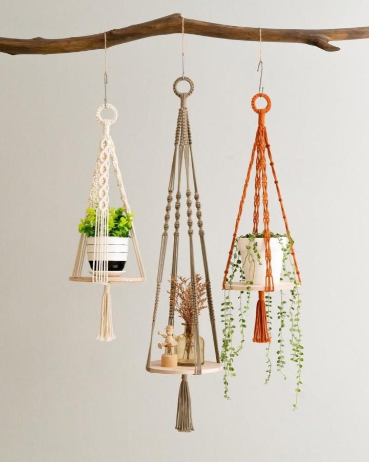 Cream, taupe, and rust hanging planters from @beandaikon. Leafy plants rest on wooden shelves beneath a rustic branch.