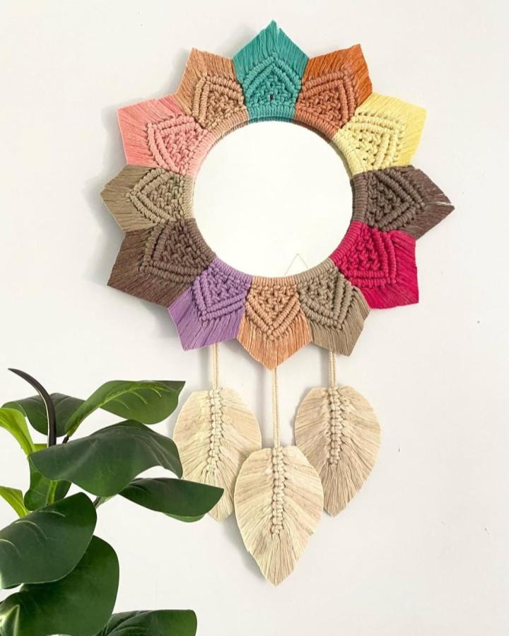 Multicolored macrame wall mirror from @rumahmacramee. Fringed petals and cream leaf tassels brighten a botanical corner.