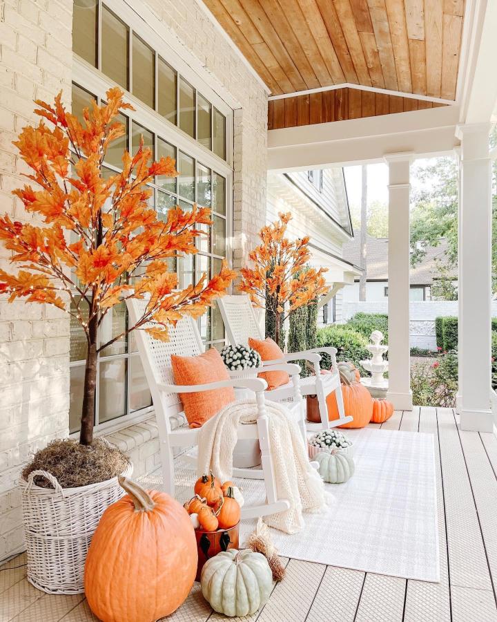 Orange faux-leaf trees and pumpkins on a white porch from @kathrynpackard. Cozy autumn seating with woven baskets.