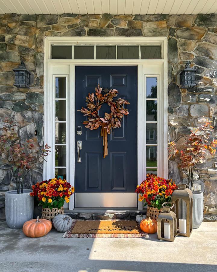 Warm-toned stone entryway from @karvicdesigns. Navy door, leafy wreath, mums, and pumpkins suit outdoor fall decor ideas.