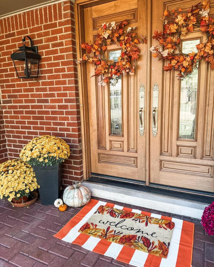 Warm autumn wreaths and mums entryway from @hillandhearthhome. Double wooden doors welcome guests with pumpkins and a fall mat.