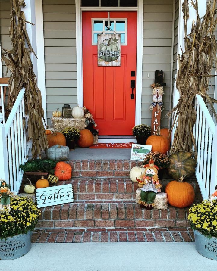 Red-orange farmhouse porch from @kraab04. Cornstalks, pumpkins, mums, hay bales, and a scarecrow welcome guests.