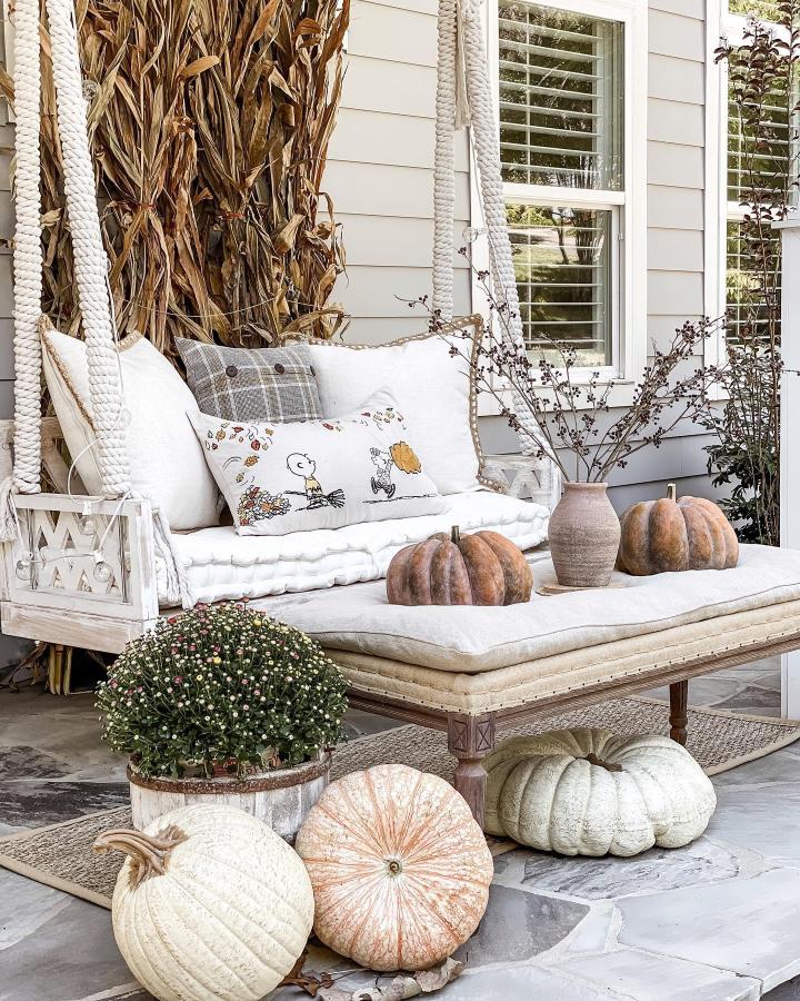 Neutral rope swing from @velveteenandgrace. Ivory cushions, pumpkins, dried corn stalks, and a flowering mum decorate the porch.