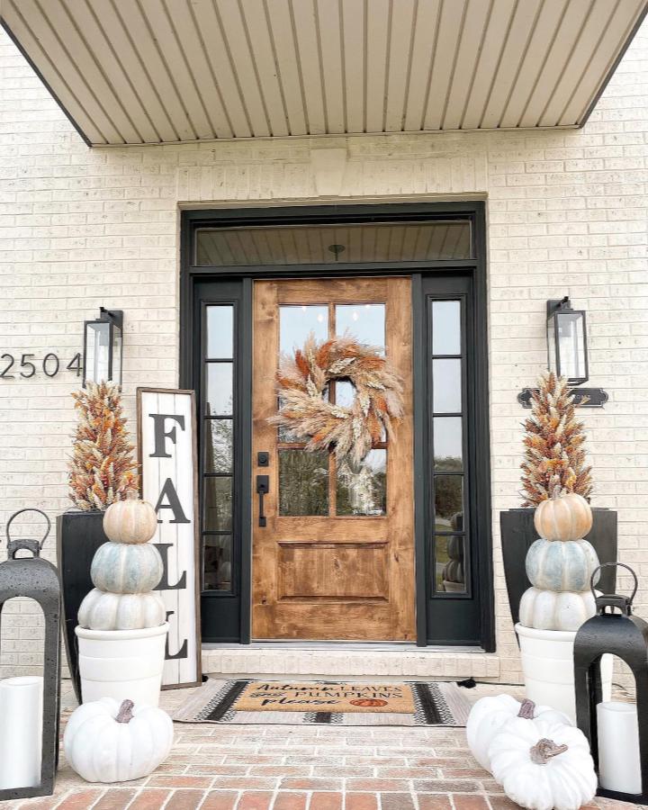 White-and-rust-toned fall porch from @homeonlickingcreek. Stacked pumpkins, wheat planters, wreath, and lanterns frame the door.