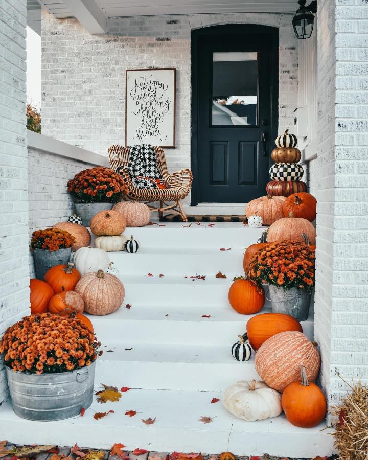 White brick fall porch with orange mums, pumpkins, wicker rocker, and checker accents from @kindredvintage. Cozy layered entryway.