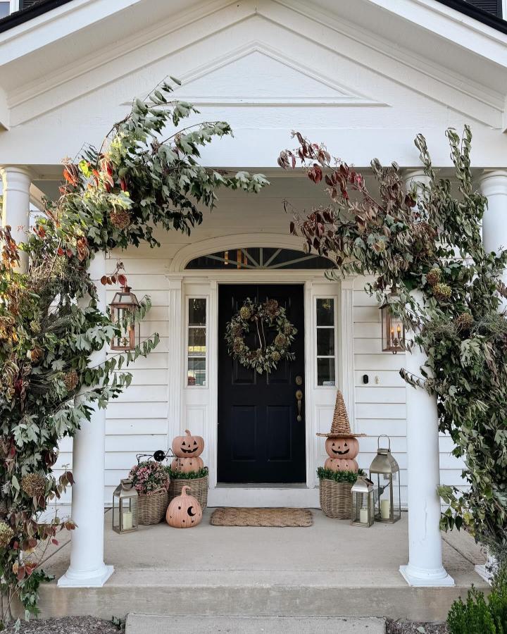 Greenery-framed white porch from @millyandomalleymarket. small fall porch decor ideas with black door, pumpkins, wicker baskets, and lanterns.