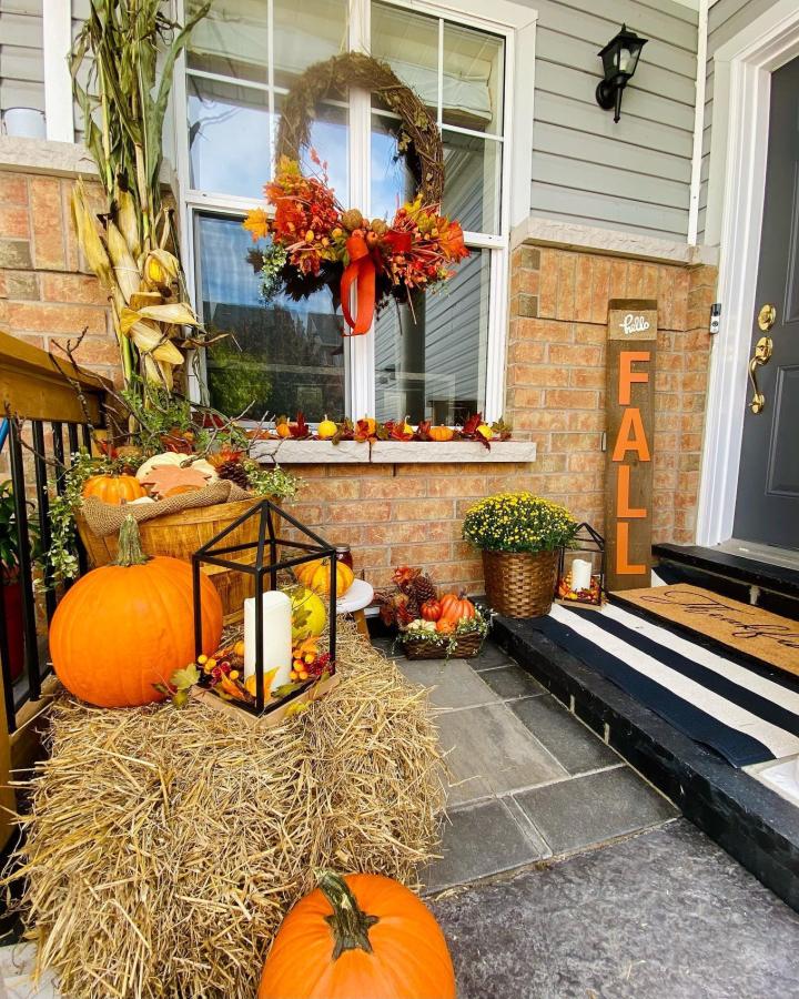 Rustic orange pumpkin porch display from @cilthestager. Hay bales, lanterns, mums, and a fall wreath brighten the compact entry.