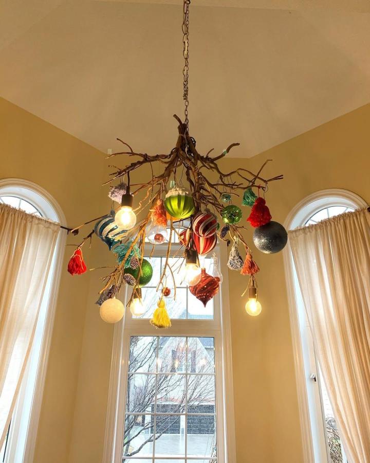 Rustic colorful branch chandelier from @inamullumani. Handmade ornaments and glowing bulbs hang above a bright window.