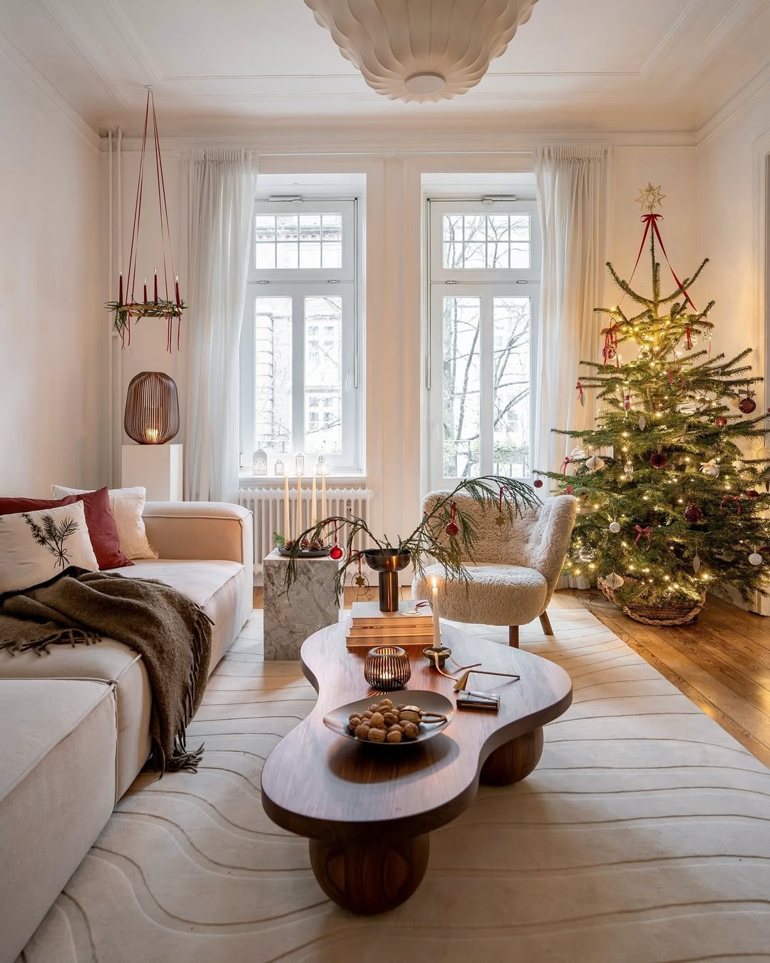 Neutral apartment living room with glowing Christmas tree, red ribbons, greenery, and cozy minimalist holiday decor