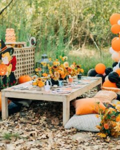 backyard halloween party ideas with rustic table, orange flowers, pumpkins, cushions, and balloon garland