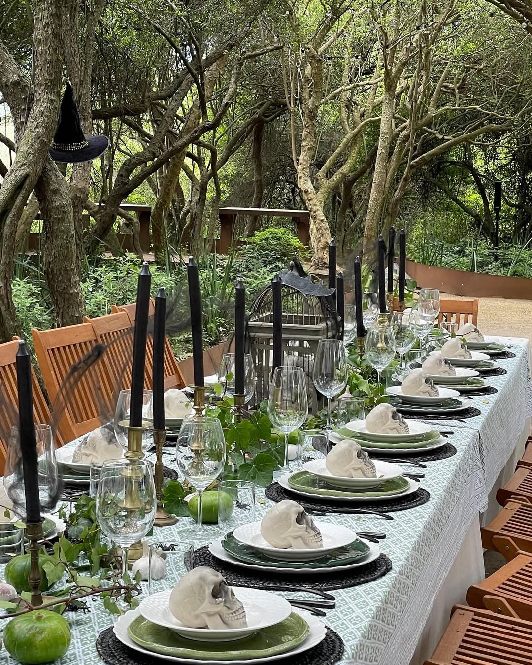 Elegant gothic Halloween tablescape with skull place settings, black candles, ivy, and a witch hat in a wooded garden