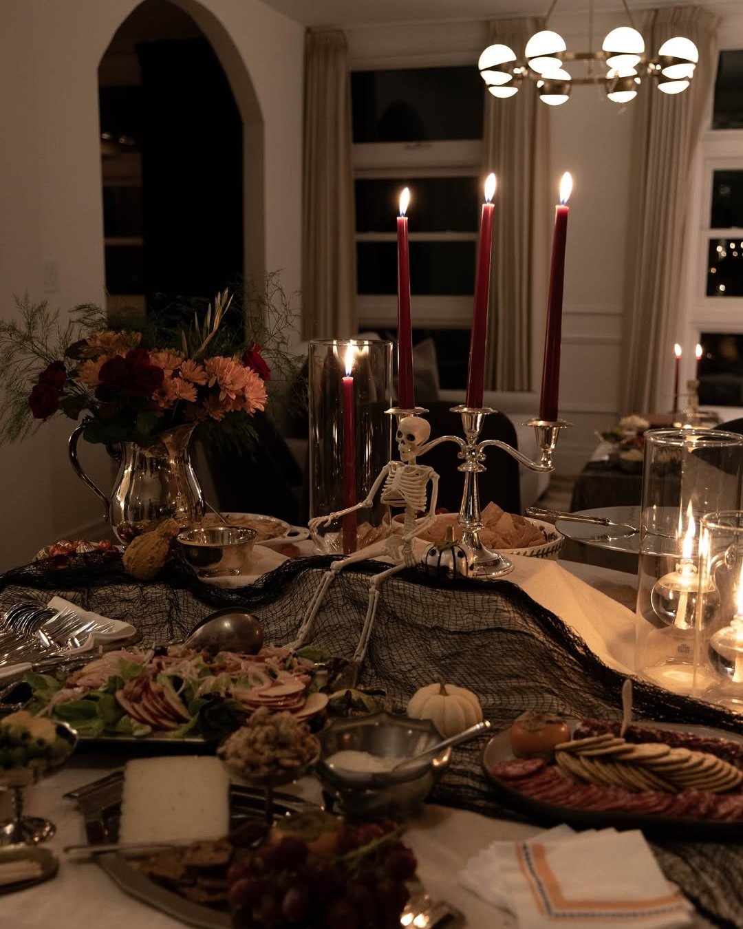 Alt text: Haunted dinner table with skeleton centerpiece, red candles, black gauze, flowers, pumpkins, and grazing boards