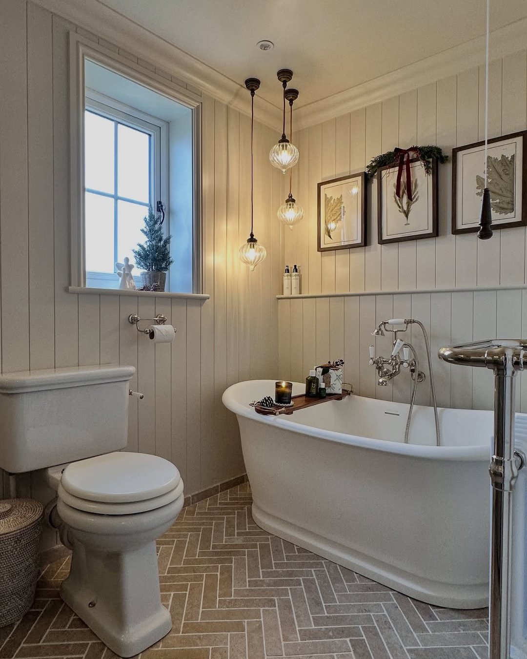 Softly lit festive bathroom with greenery, red ribbon, botanical art, and a clawfoot tub