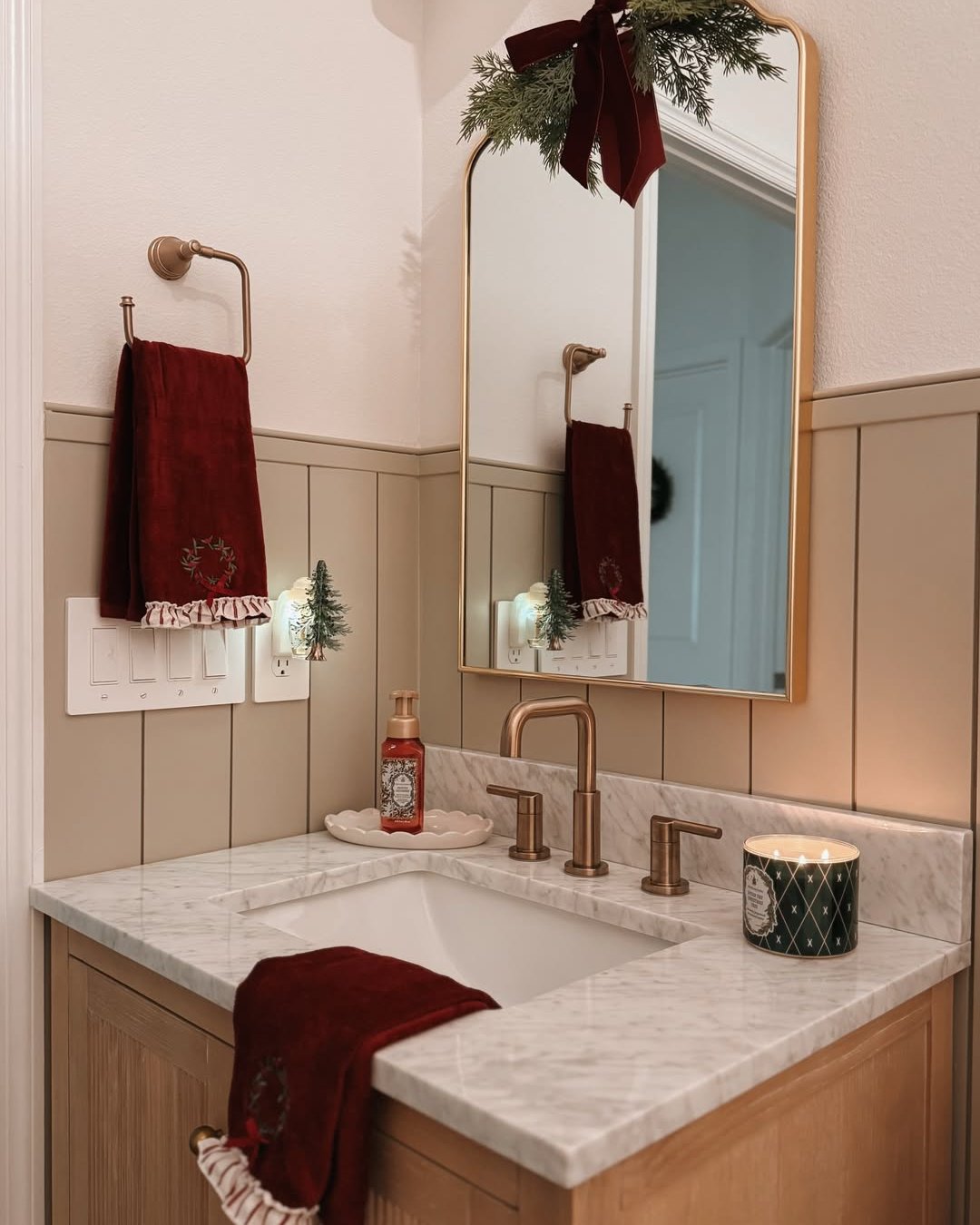 Burgundy towels, evergreen mirror garland, candle, and miniature trees create festive bathroom styling