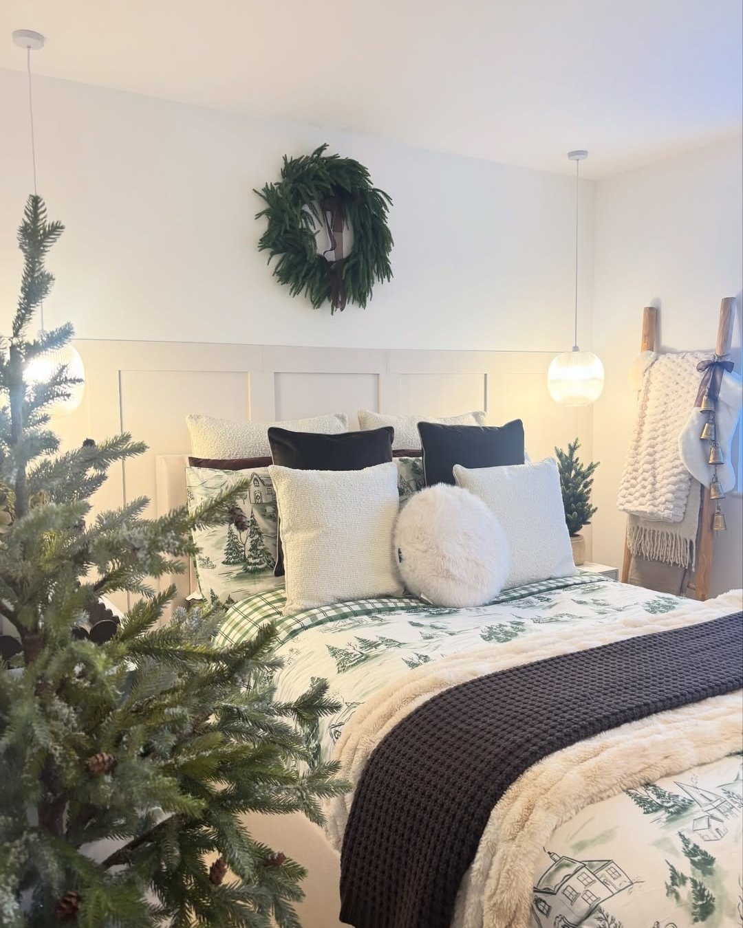 Cozy holiday bedroom with evergreen wreath, tree branches, plaid bedding, layered pillows, and a chunky knit runner