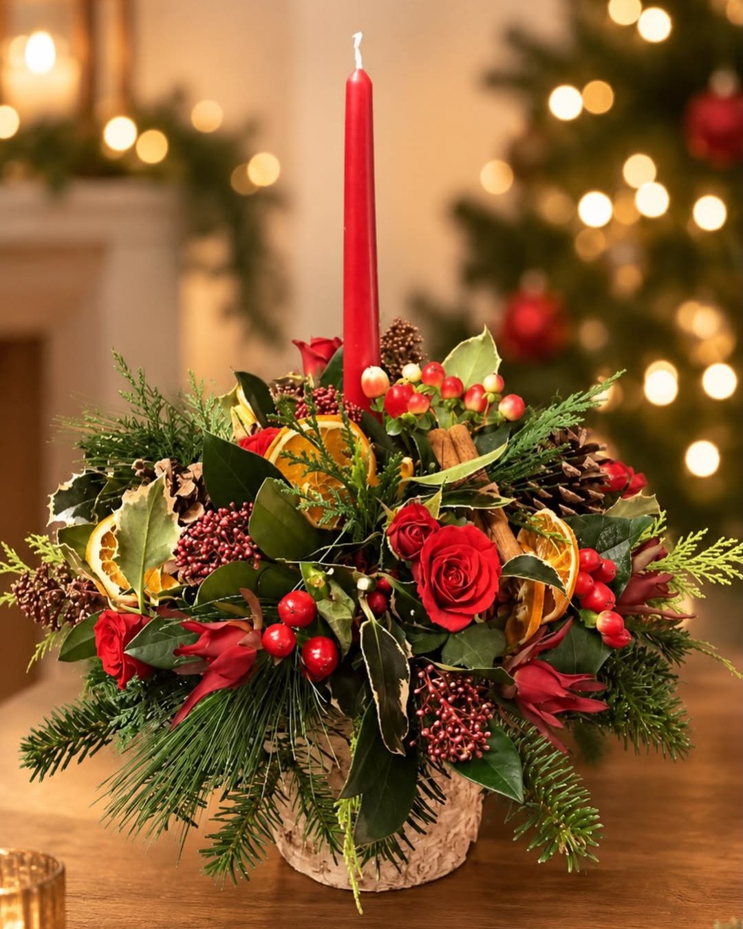 Red candle evergreen centerpiece with roses, berries, citrus, pinecones, and festive greenery