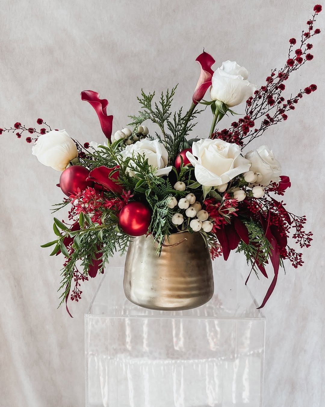 White roses, burgundy calla lilies, red ornaments, and winter greenery in a metallic holiday centerpiece