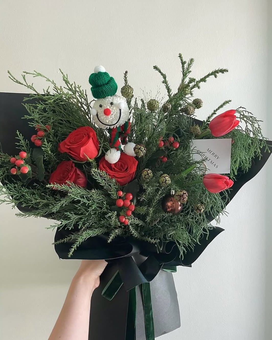 Red roses, evergreen branches, berries, tulips, and a smiling snowman create a festive holiday bouquet
