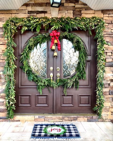Lush greenery, wreaths, red ribbon, golden bells, and a plaid doormat decorate a double entry door