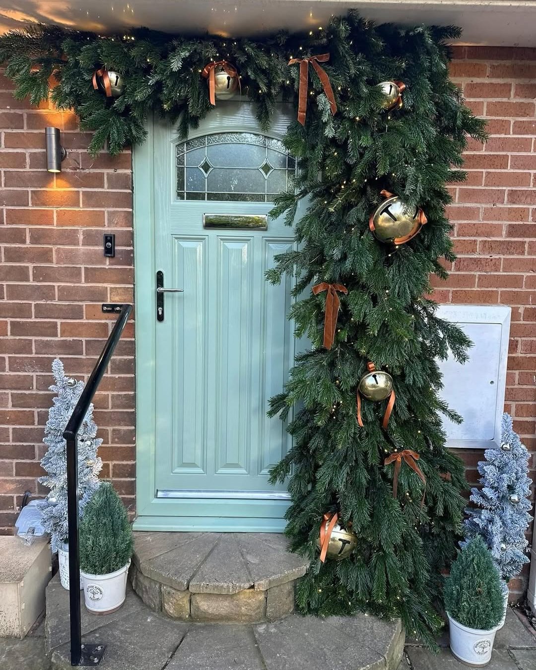 Evergreen bell garland framing a mint green door with glowing lights and festive potted trees