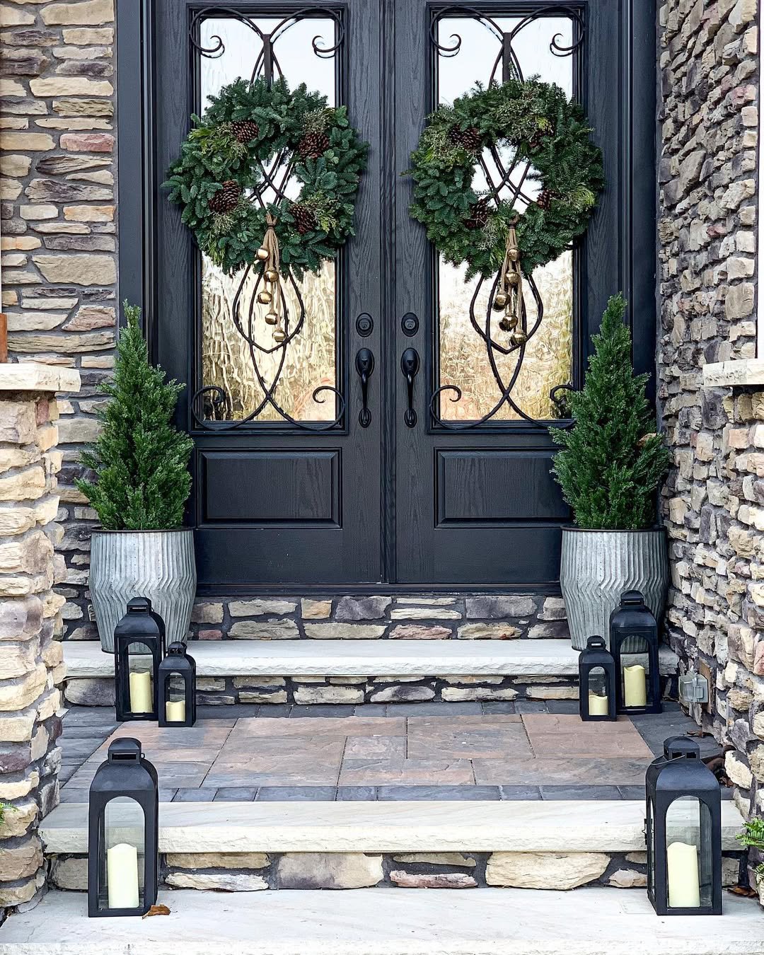 Twin evergreen wreaths, potted trees, and black lanterns decorate a stone entryway for Christmas