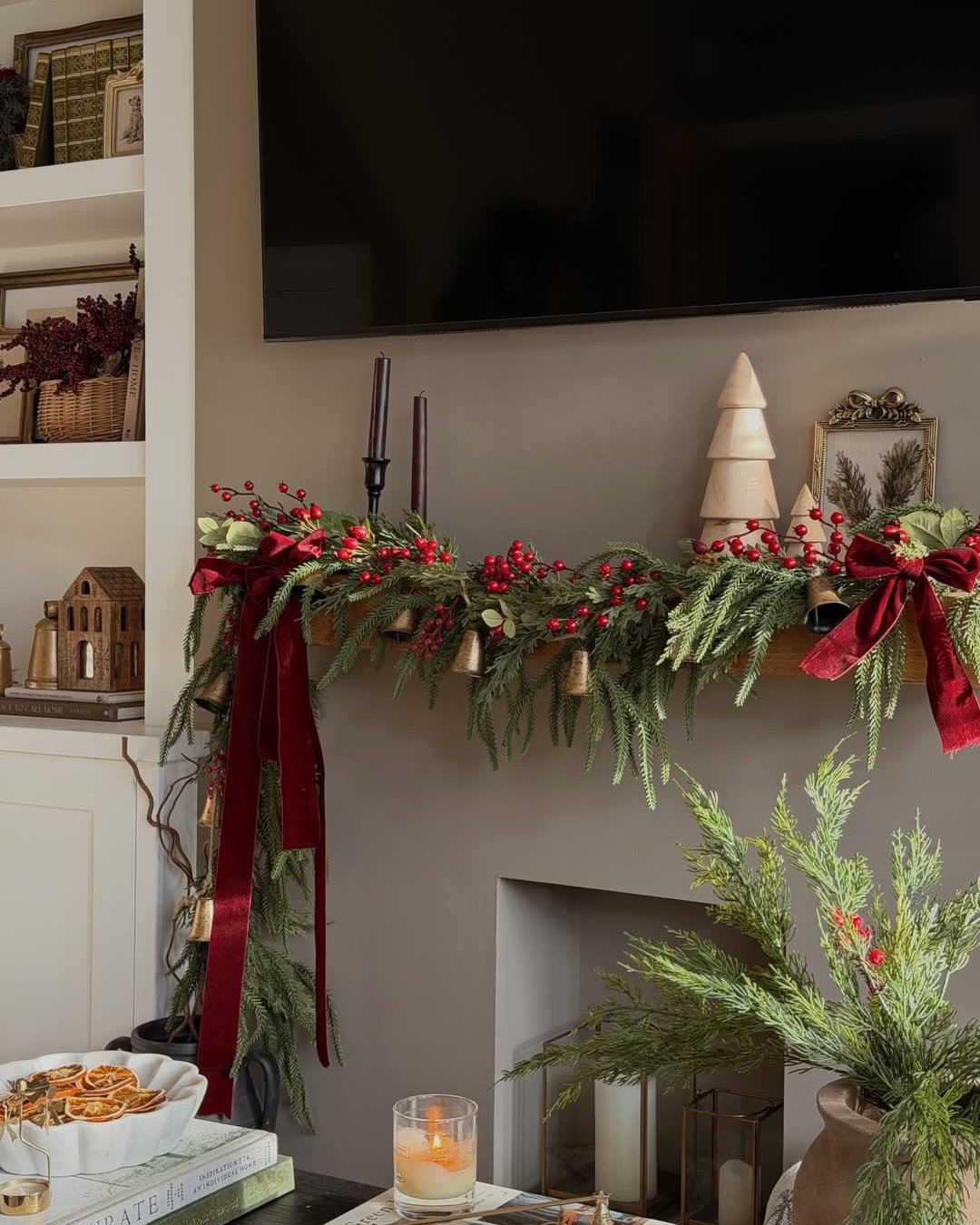 Lush evergreen mantel garland with red berries, velvet bows, and brass bells above a holiday fireplace