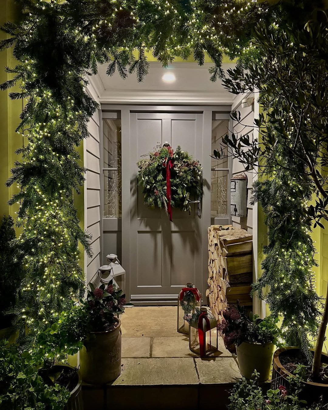 Lit evergreen doorway garland with glowing lights, wreath, red ribbon, lanterns, and potted greenery