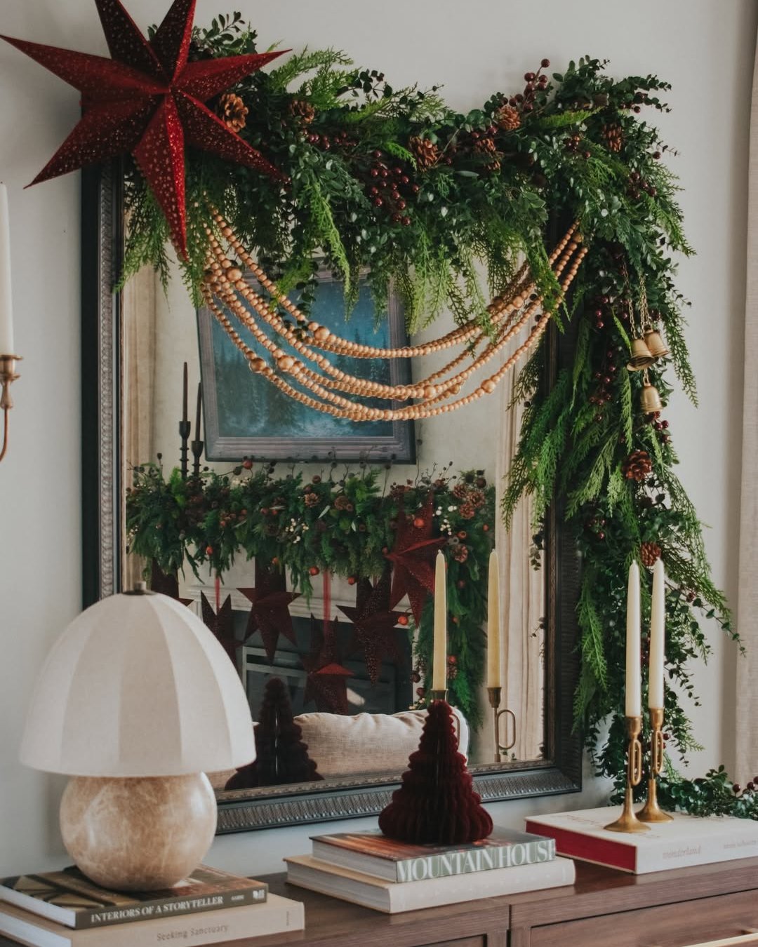 Lush Christmas greenery garland with wooden bead swags, pinecones, berries, bells, and a red star above a mirror