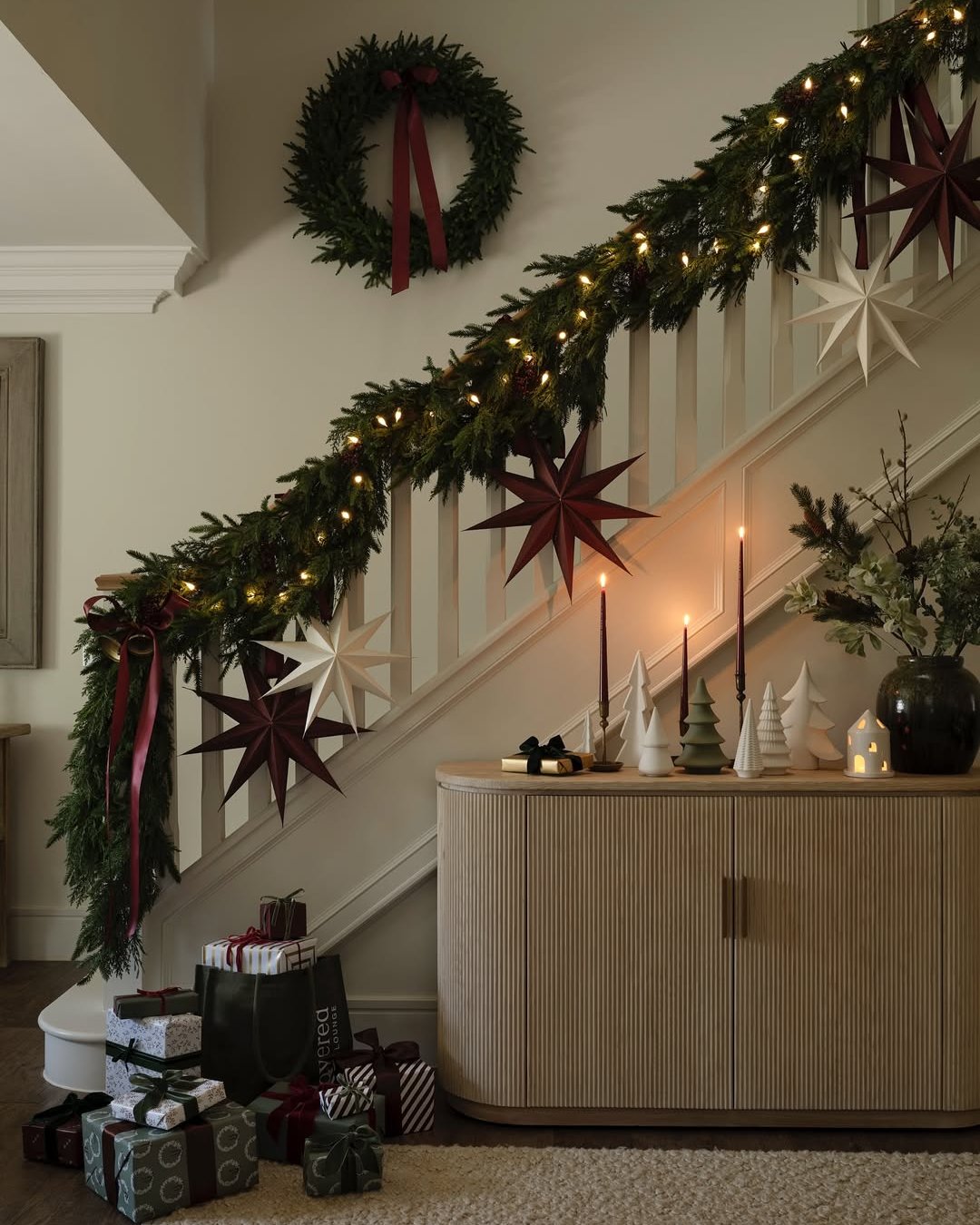 Evergreen staircase garland with glowing lights, paper stars, burgundy ribbons, and matching holiday wreath
