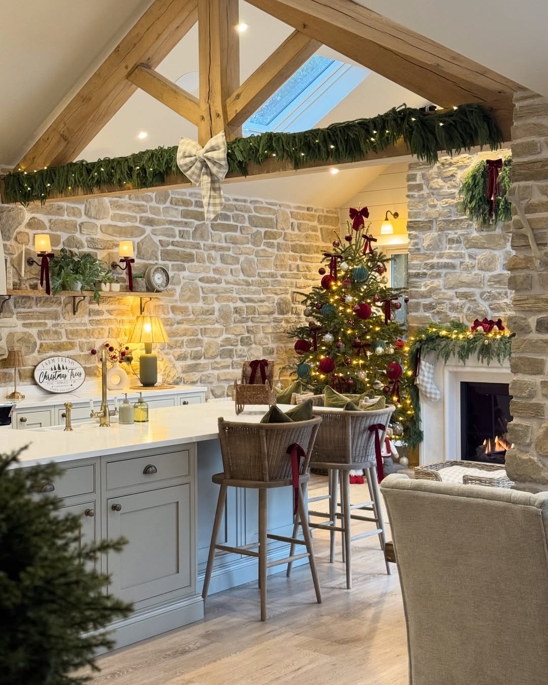 Rustic kitchen with glowing evergreen garland, checked bows, Christmas tree, and stone fireplace