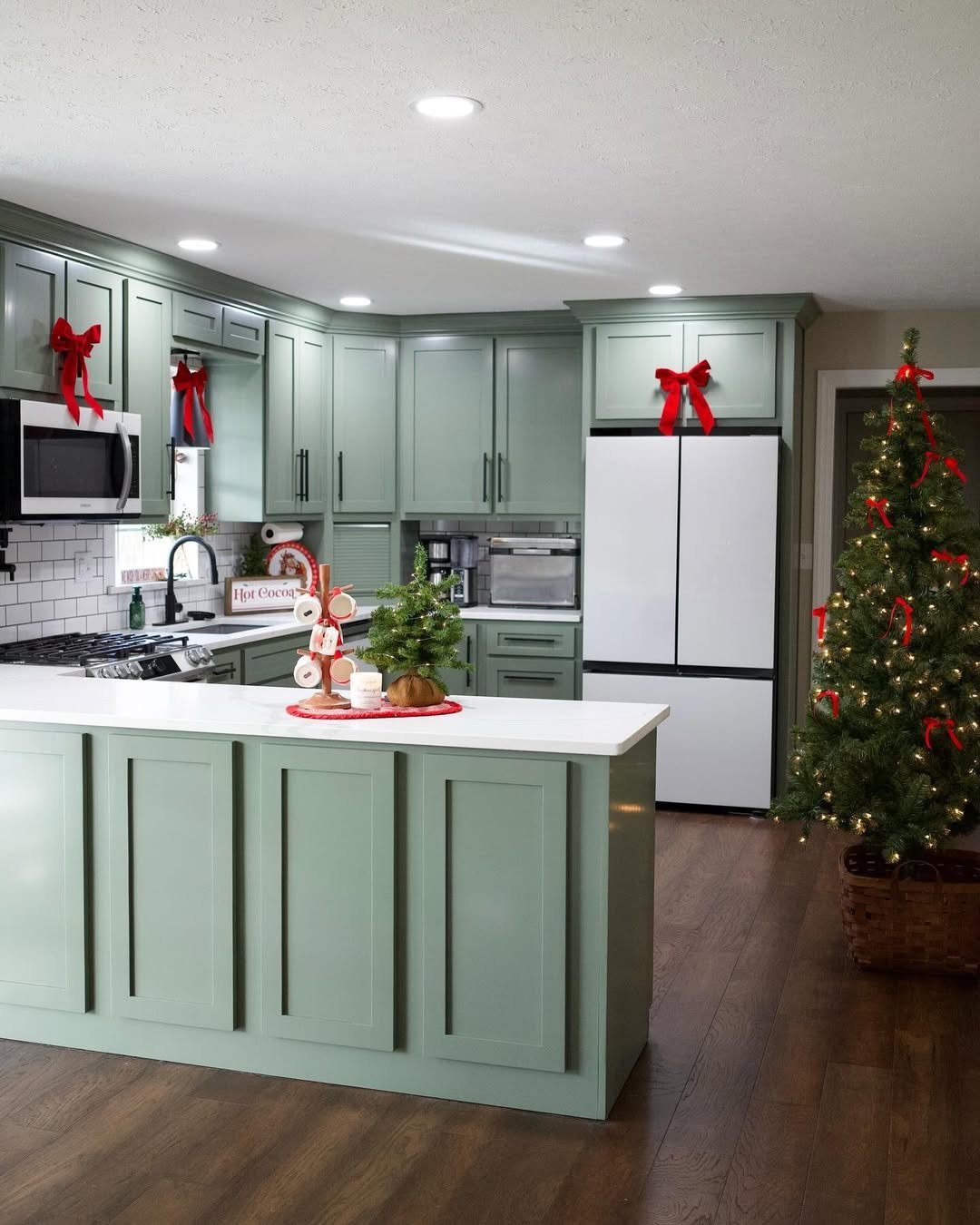 Green kitchen cabinets with red bows, a hot cocoa display, and a lit Christmas tree