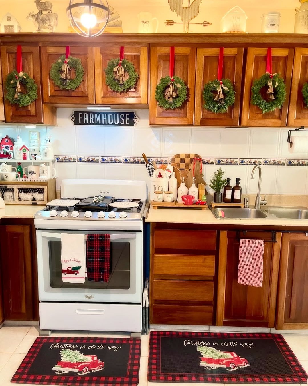 Cozy farmhouse Christmas kitchen with cabinet wreaths, bells, plaid accents, and festive greenery