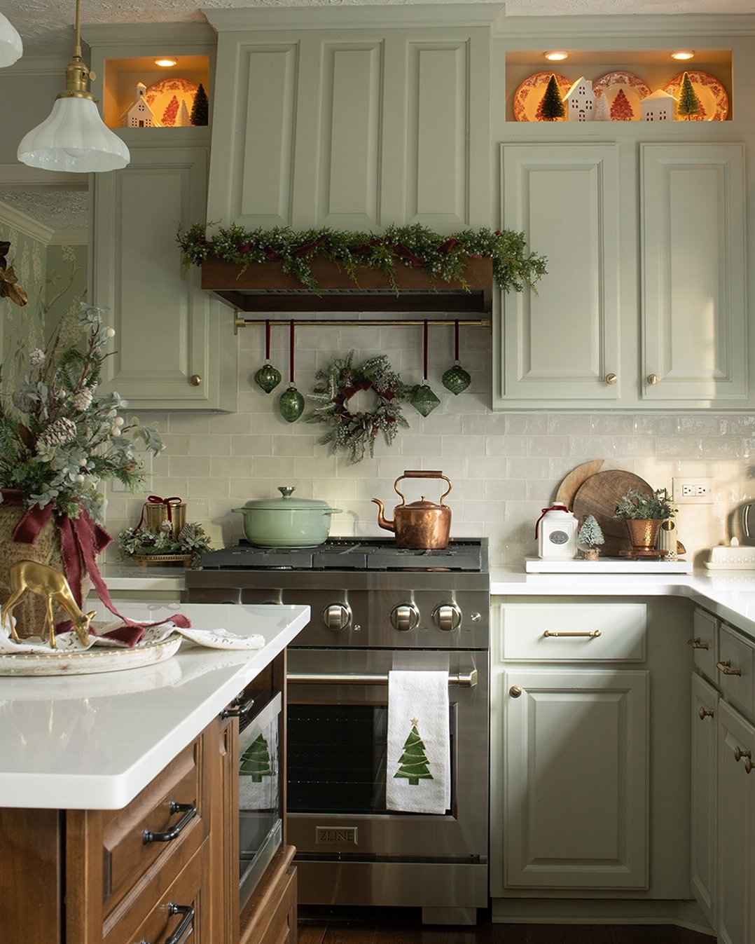 Warm Christmas kitchen with evergreen garland, hanging ornaments, village houses, and festive wreath