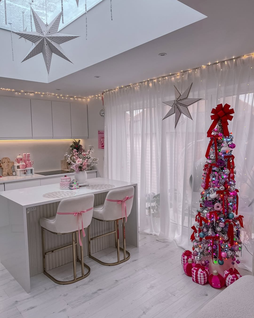Festive pink Christmas kitchen with ribbon-wrapped tree, glowing lights, pastel ornaments, and oversized hanging stars