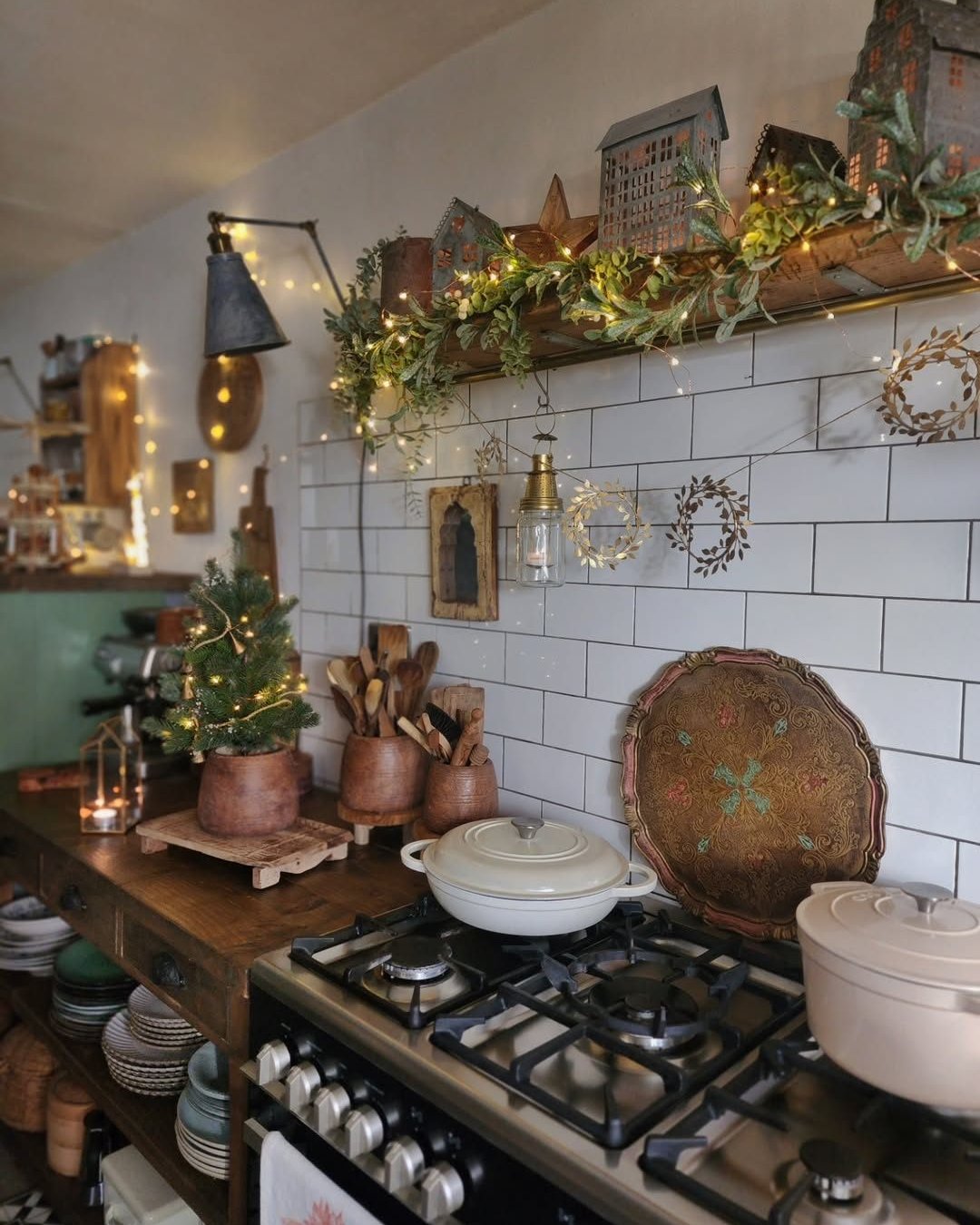 Rustic Christmas kitchen with glowing garland, miniature village houses, evergreen tree, white tile, and copper-toned accents