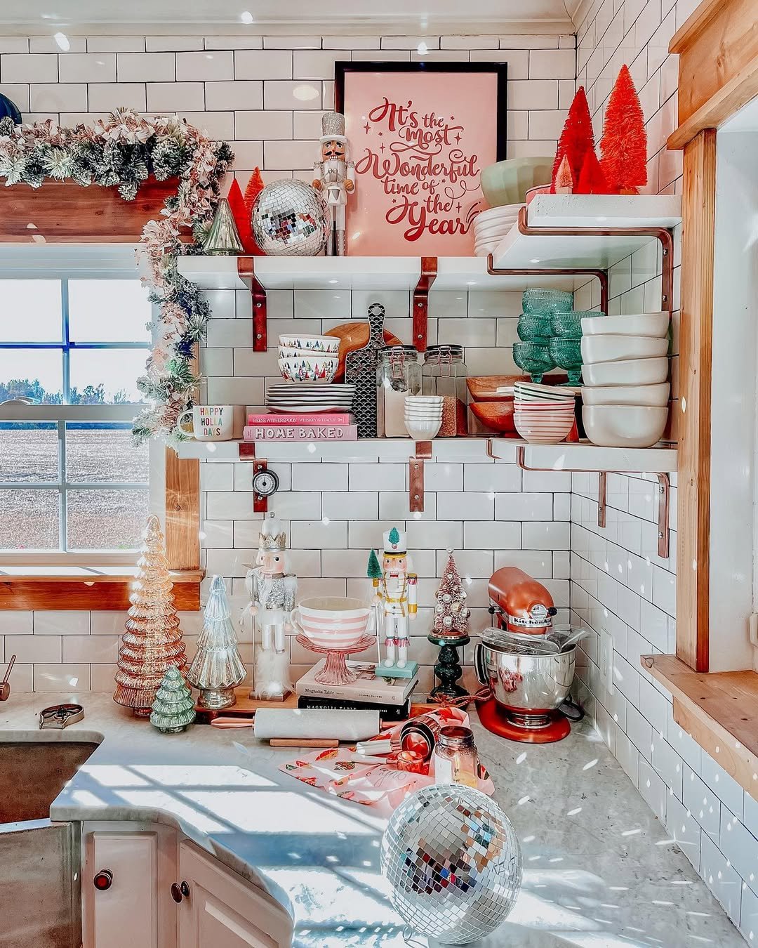 White kitchen shelves decorated with Christmas garland, nutcrackers, bottle-brush trees, red dishes, and disco balls