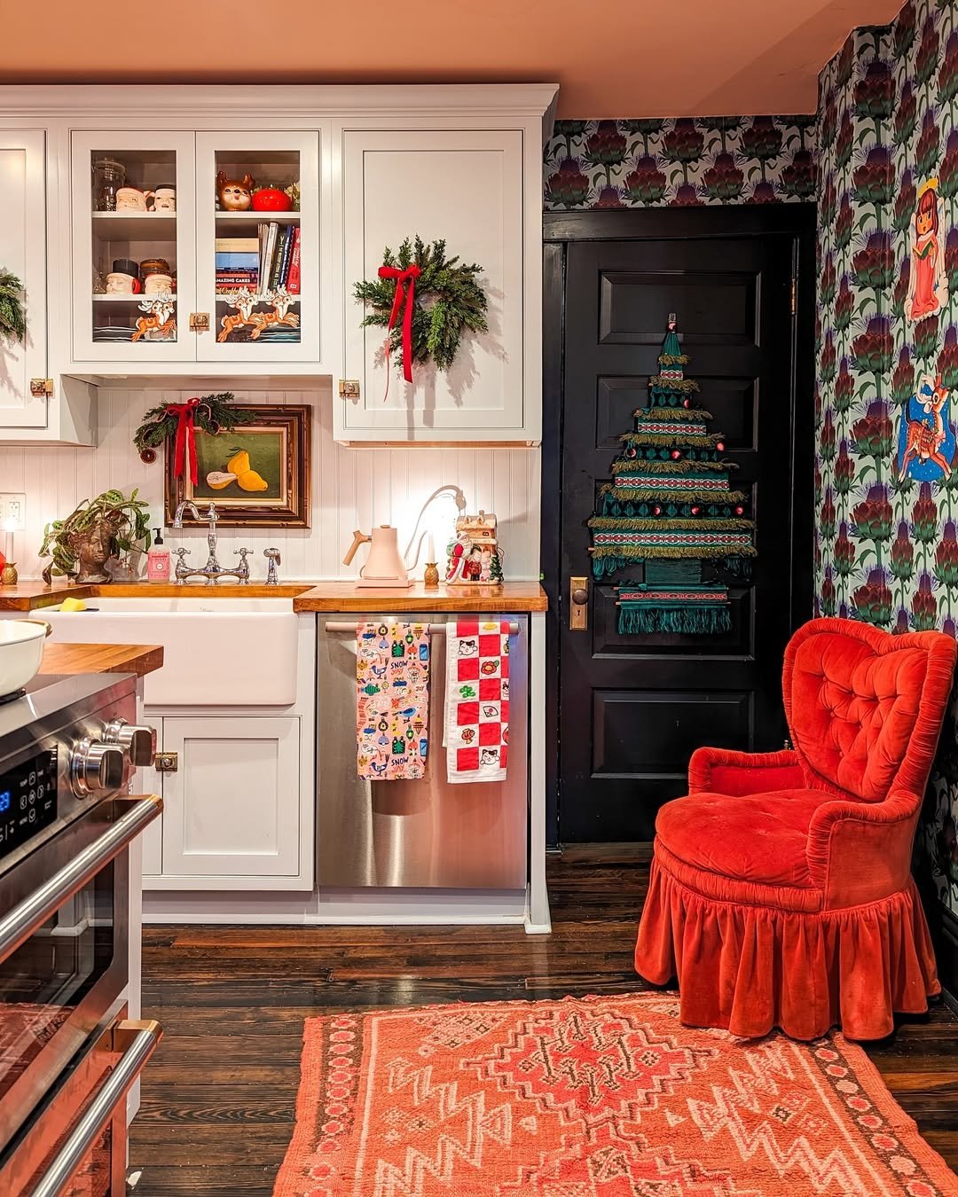 Festive kitchen with cabinet greenery, red ribbons, textile tree, patterned wallpaper, and holiday towels