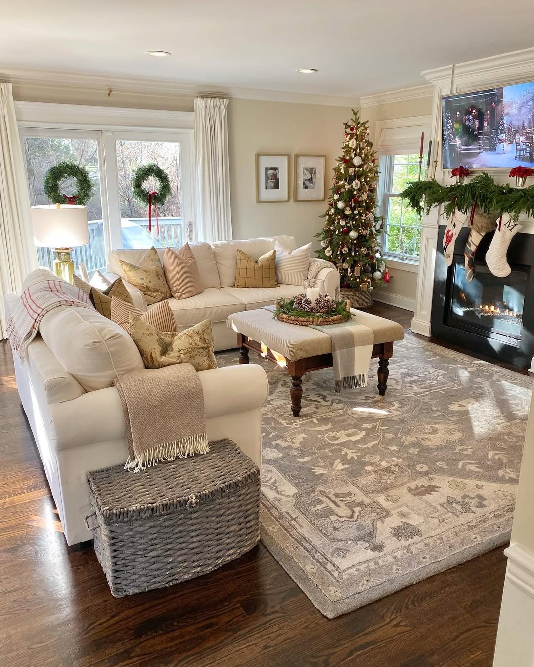 Warm neutral Christmas living room with decorated tree, window wreaths, fireplace garland, stockings, and cozy furnishings
