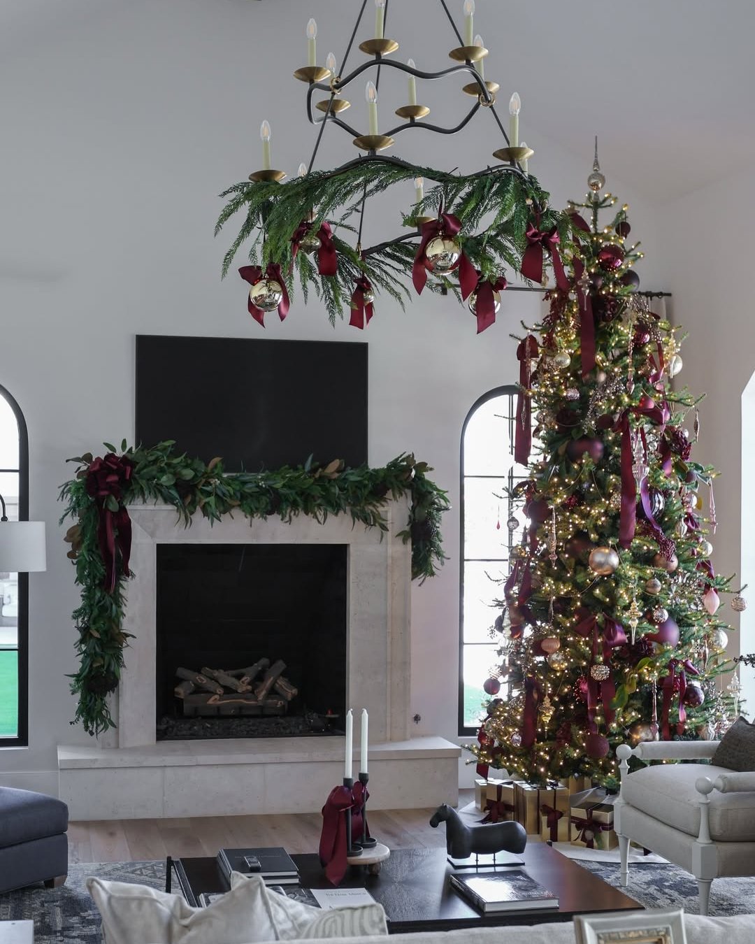 Evergreen chandelier, burgundy ribbons, decorated tree, and glowing fireplace in an elegant holiday living room