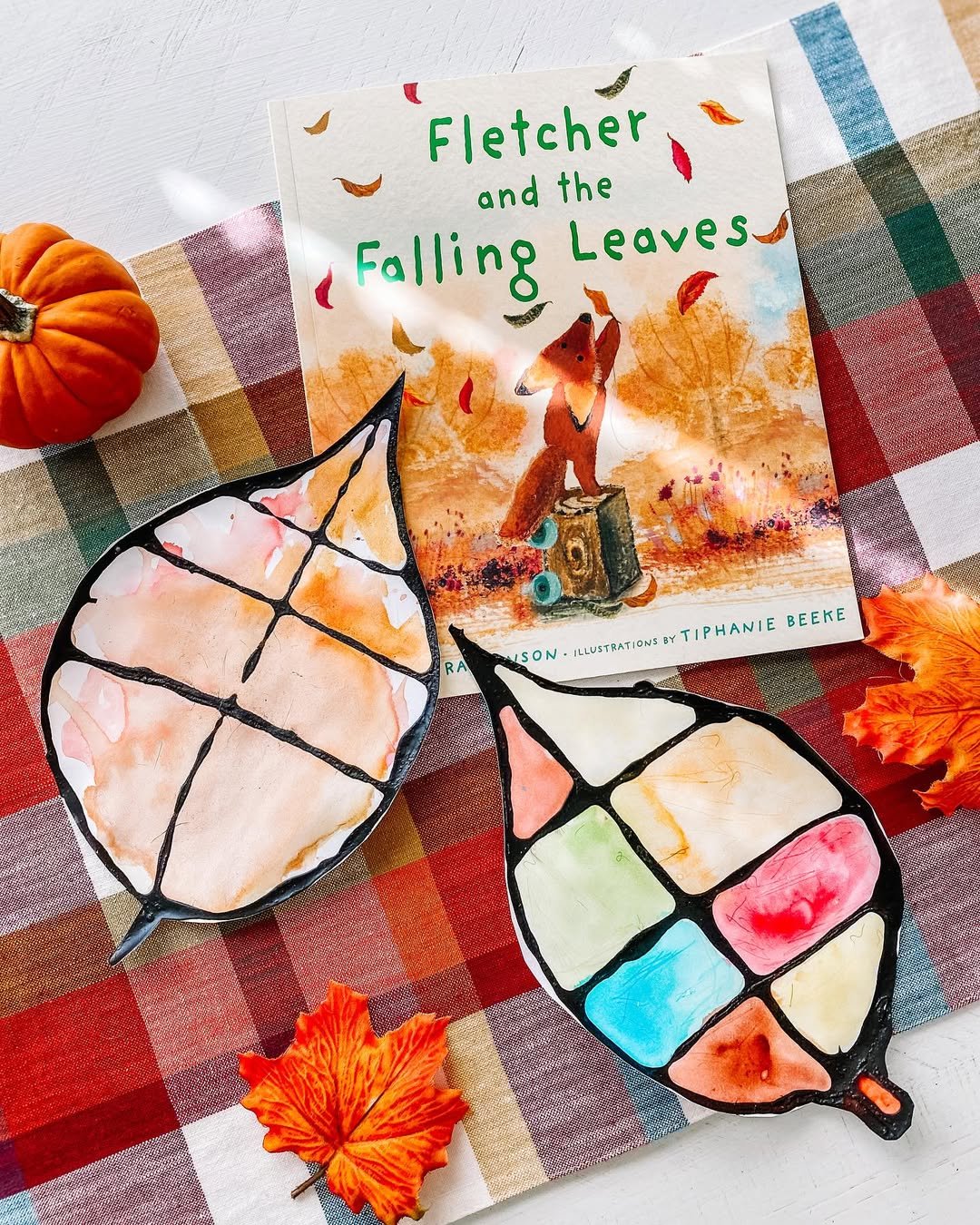 Watercolor leaf suncatchers with colorful sections beside a fall storybook and pumpkins