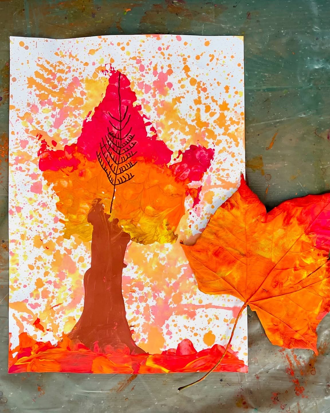 Children's autumn art with a colorful painted tree, textured leaves, and orange splatters on white paper