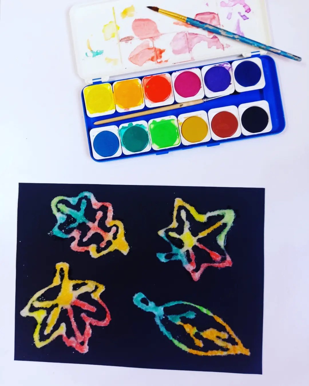 Colorful salt-painted leaves on black paper with watercolor supplies for fall leaf crafts for kids