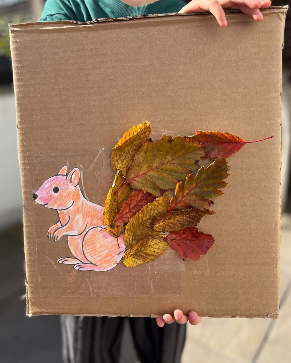 A colorful fox drawing with layered autumn leaves forming its bushy tail on cardboard