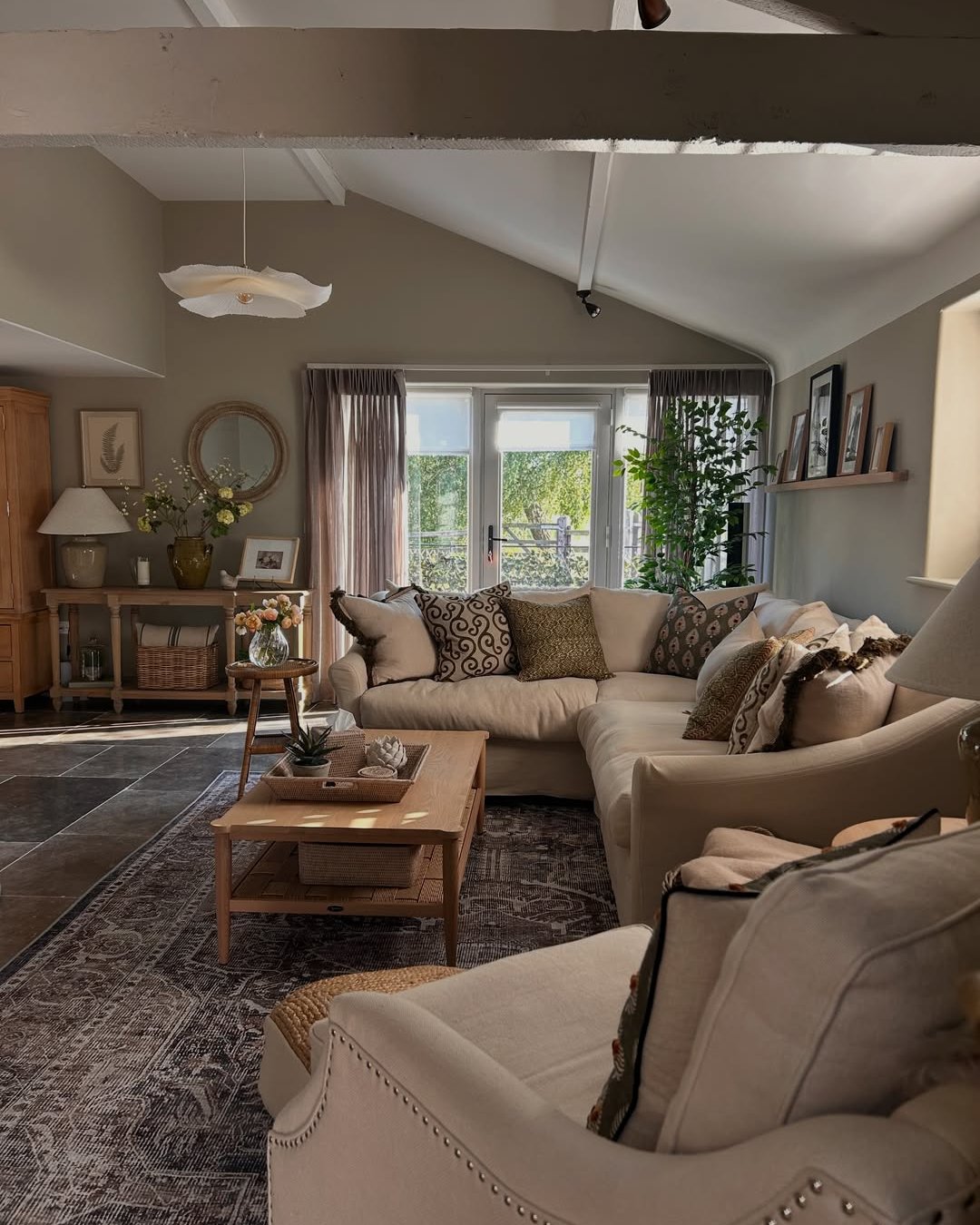 Cozy winter lounge with layered cushions, warm neutrals, patterned rug, wood accents, and leafy greenery